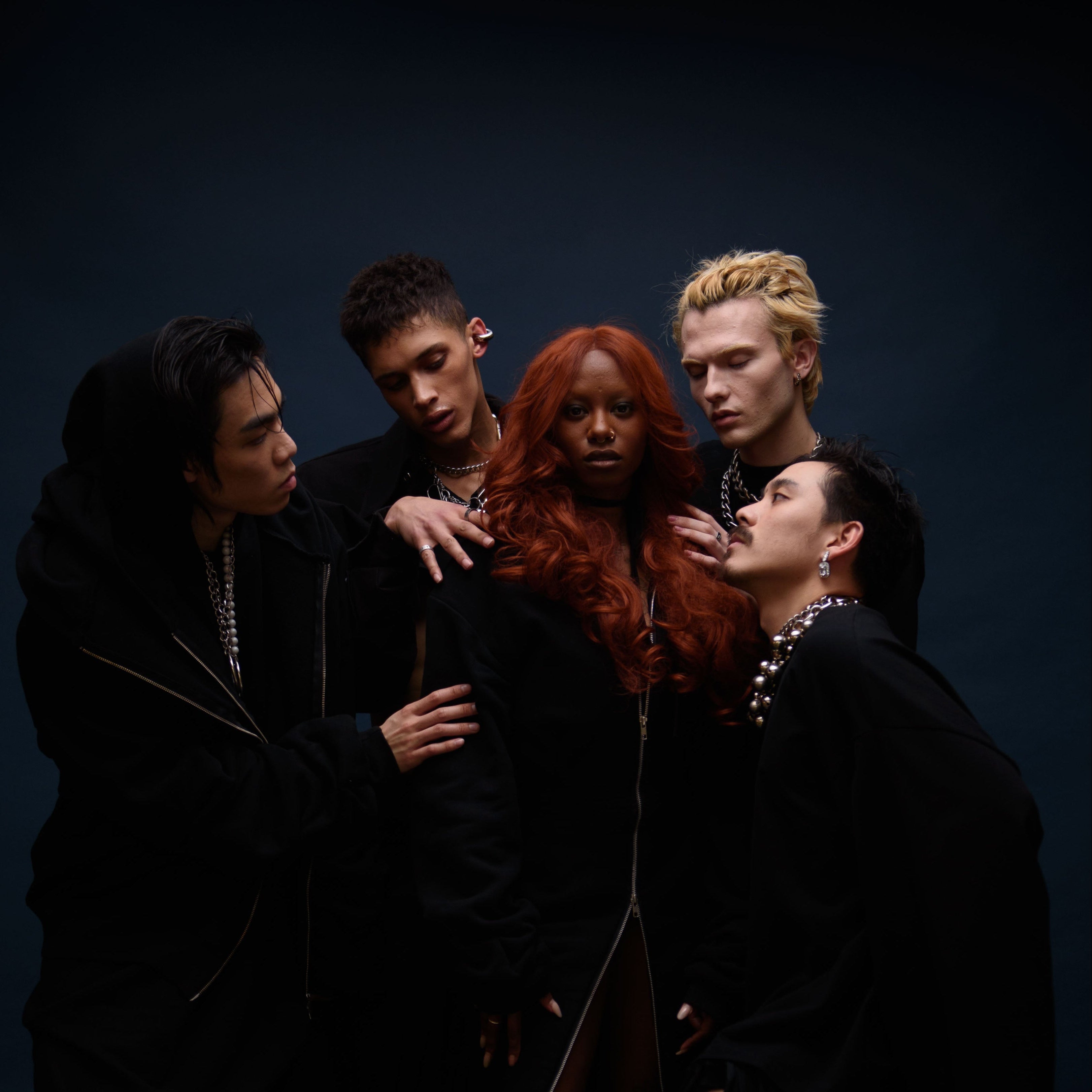 Five people in black clothing posed together against a dark blue studio background. The image is rotated sideways, and the lighting is low-key with strong shadows.